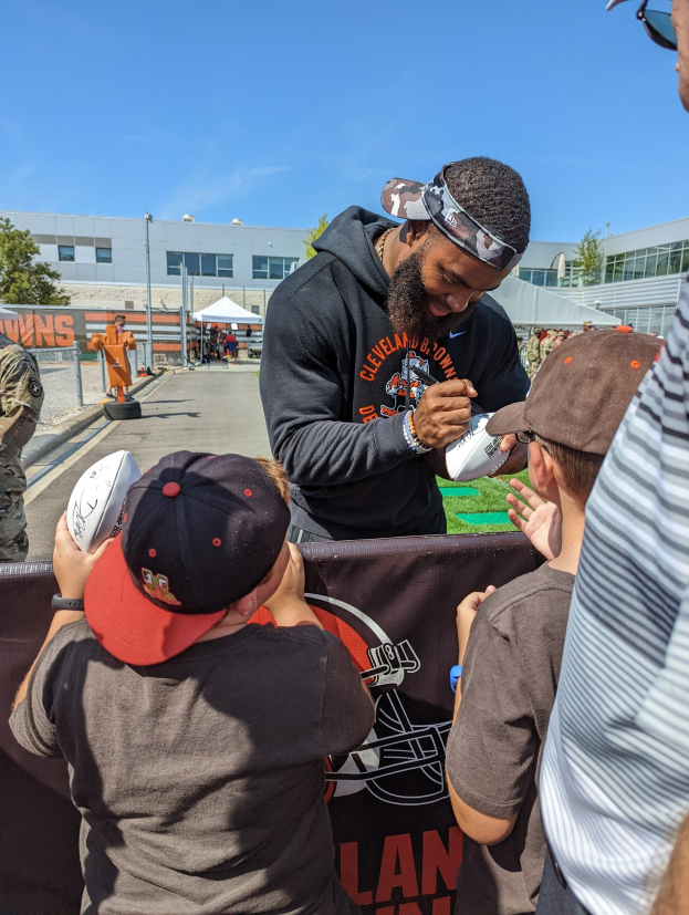 Cleveland Browns Wide Receiver Odell Beckham Jr. signiert Autogramme für Fans auf dem Trainingsgelände des Teams, umgeben von Menschen mit Kappen, einem Banner und Gebäuden im Hintergrund bei bewölktem Himmel.