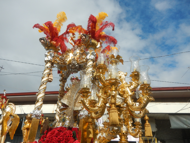 Ein prächtiger goldener und roter Festwagen, geschmückt mit Blumen und anderen Dekorationsartikeln bei einem Karnevalsumzug, mit einem Gebäude, Strommasten mit Drähten und einem bewölkten Himmel im Hintergrund.