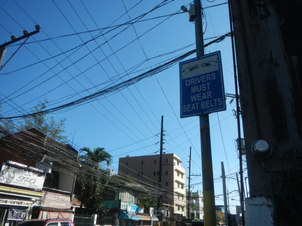 Eine Stadtstraße mit Autos, Strommasten mit Drähten, Gebäuden, Bäumen und Namensschildern, mit einem Schild an einem Strommasten, auf dem steht: "Fahrer müssen Sicherheitsgurte tragen".