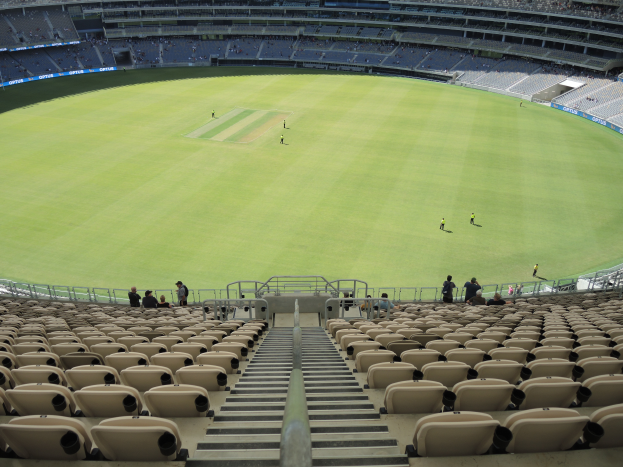 Ein großes Stadion voller Zuschauer bei einem Cricket-Spiel, mit grünem Rasen, Stufen, Geländern und Bannern mit Text.