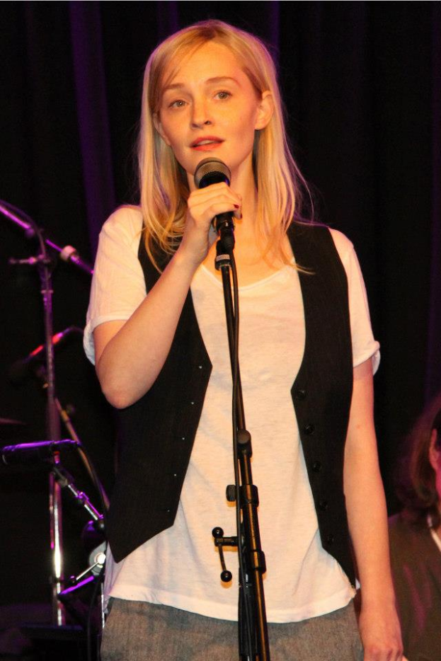 Eine Frau in einem wei├čen T-Shirt und einer schwarzen Jacke steht auf der B├╝hne und h├Ąlt ein Mikrofon in der Hand, mit einer anderen Person rechts von ihr und Musikinstrumenten im Hintergrund.