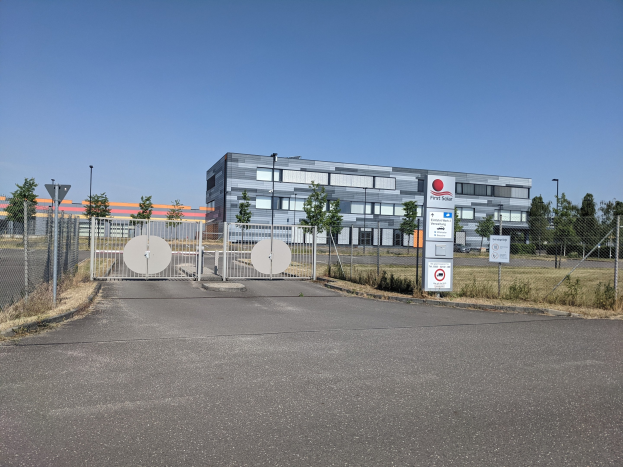 Großes industrielles Gebäude mit der Aufschrift "First Solar" und zahlreichen Fenstern, umgeben von einem Metallzaun, Bäumen, Pflanzen, Gras, Informationsschildern, Straßenlaternen und einem Weg unter einem bewölkten Himmel.