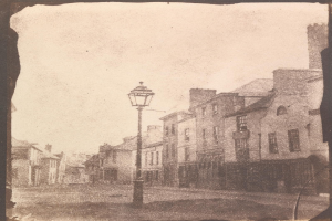 Altes Schwarz-Weiß-Foto einer Straßenlaterne zwischen Gebäuden, die eine Stadtstraße säumen, unter einem klaren Himmel.