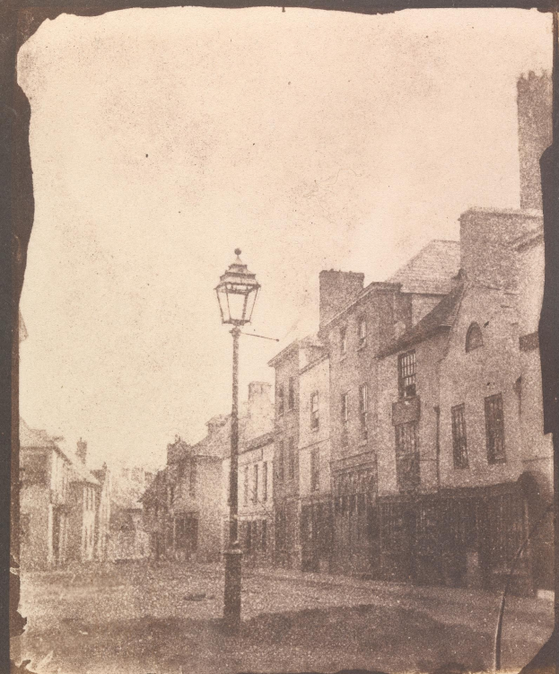 Altes Schwarz-Weiß-Foto einer Straßenlaterne zwischen Gebäuden, die eine Stadtstraße säumen, unter einem klaren Himmel.