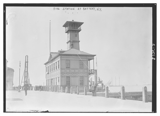 Schwarzes und weißes Foto einer Feuerwache in Battery, New York, mit Menschen, Pfählen, einem Zaun, Booten auf dem Wasser und einem bewölkten Himmel.