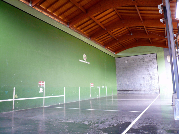 Gro├če Indoor-Tennishalle mit grünen W├Ąnden, einer h├Âlzernen Decke und einer Stange auf der rechten Seite sowie deckenumlaufenden Leuchten.