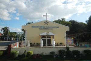 Eine unabhängige Kirche mit einem Kreuz vorne, umgeben von einem Zaun, Statuen, Pflanzen, Gras, einem Weg, Bäumen, Pfählen, Drähten, einem nahen Haus und einem bewölkten Himmel.