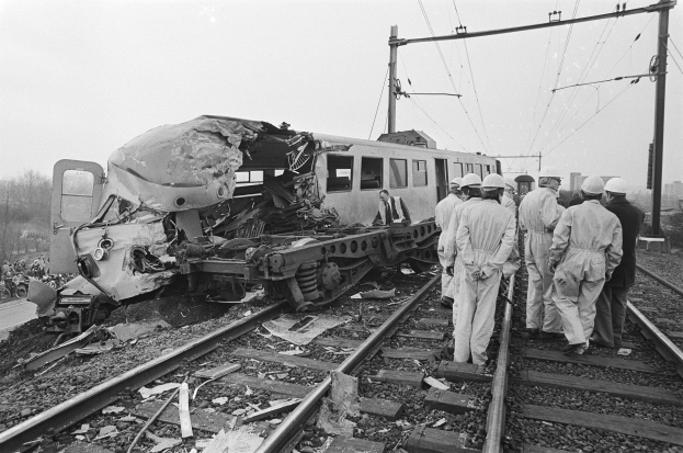 Eine Schwarz-Weiß-Szene eines Zugunglücks mit zwei Zügen auf den Schienen, umgeben von Menschen in Helmen, mit Strommasten, Bäumen, Gebäuden und einem klaren Himmel im Hintergrund.