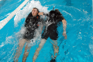 Zwei Personen in schwarzen Neoprenanzügen und Tauchausrüstung, eine mit einem Sauerstoffzylinder, führen einen Rückenschwimmzug in einem Schwimmbad durch.