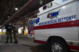 American Medical Response ambulance parked inside a building with two masked individuals in front of it, a vehicle to the left, and additional people and objects in the background.