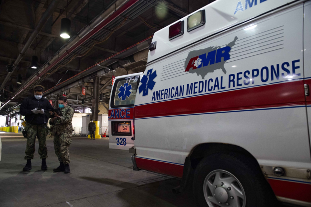 American Medical Response ambulance parked inside a building with two masked individuals in front of it, a vehicle to the left, and additional people and objects in the background.