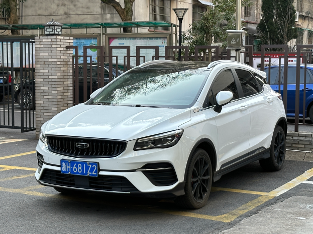 Ein weißes Volvo XC40 T5 AWD Inscription auf einem Parkplatz mit Gebäuden, Bäumen, Masten, Lichtern und anderen Objekten im Hintergrund.
