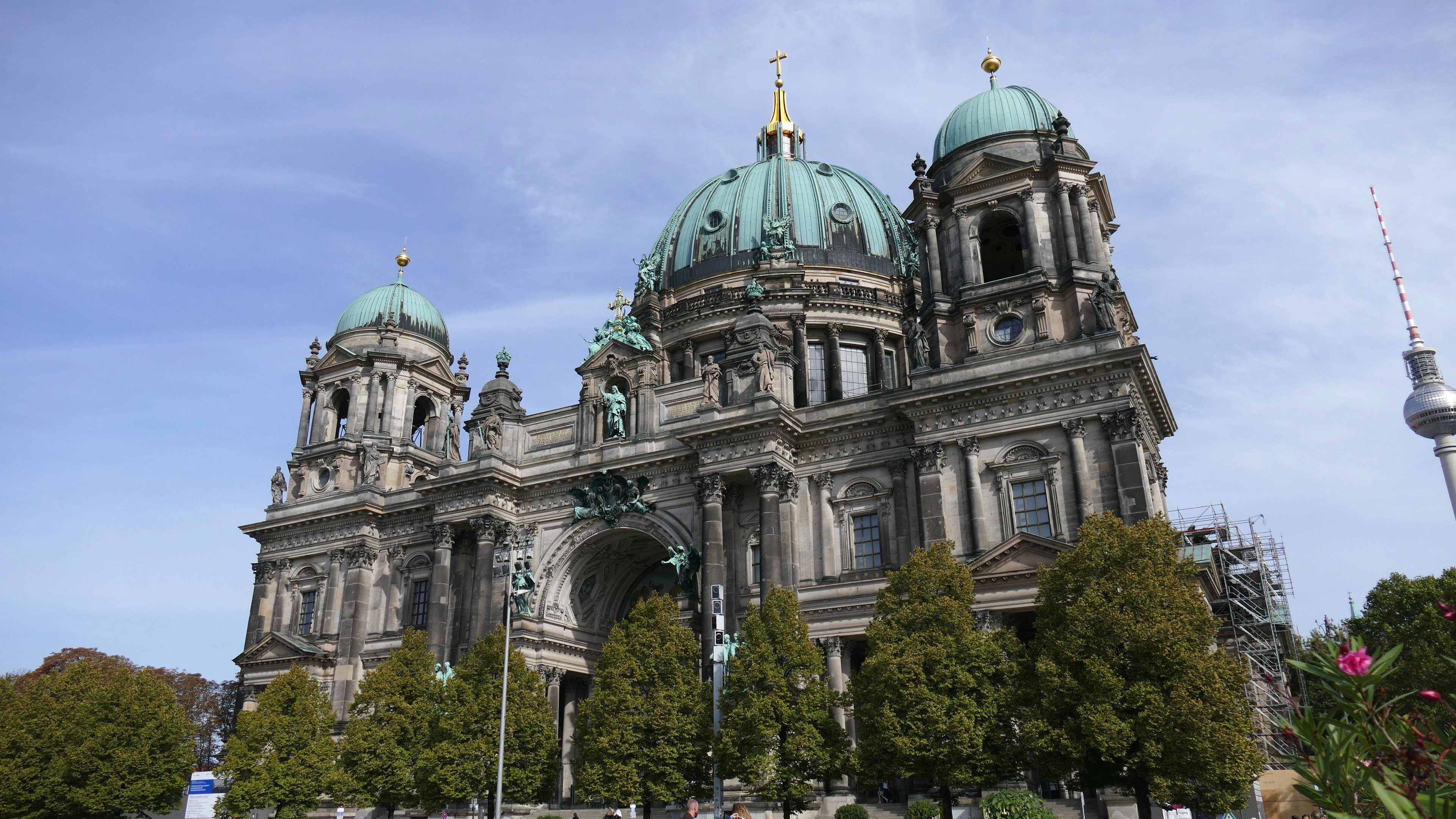 Der Berliner Dom, eine große Kathedrale mit detaillierter Architektur, steht prominent in Berlin, Deutschland, mit Menschen in der Nähe und Bäumen, die die Straße säumen, unter einem bewölkten Himmel.