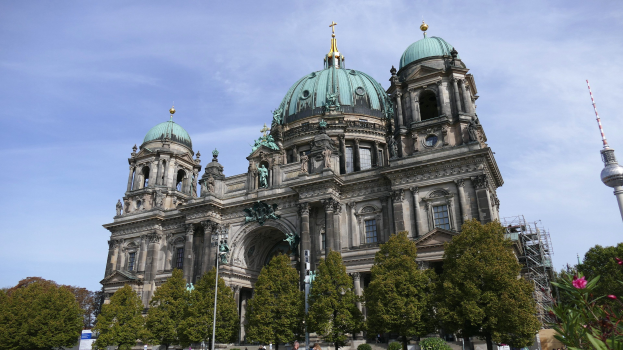 Der Berliner Dom, eine große Kathedrale mit detaillierter Architektur, steht prominent in Berlin, Deutschland, mit Menschen in der Nähe und Bäumen, die die Straße säumen, unter einem bewölkten Himmel.