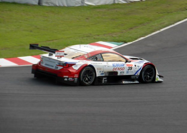Ein Sportwagen mit Text und Zahlen, der bei den 24 Stunden von Le Mans teilnimmt, fährt auf einer Rennstrecke mit Gras an beiden Seiten und einem Zaun im Hintergrund.