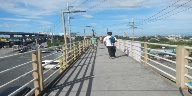 Eine Gruppe von Menschen überquert eine Fußgängerbrücke über einer vielbefahrenen Straße, mit Geländern, Strommasten und Drähten, während Fahrzeuge darunter fahren und Gebäude, Bäume und ein bewölkter Himmel im Hintergrund zu sehen sind.
