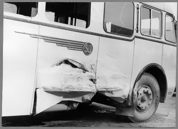Ein Schwarz-Weiß-Foto eines beschädigten Busses am Straßenrand, dessen Vorderseite eingedrückt ist, Fenster zersplittert und die Fahrertür offen steht.
