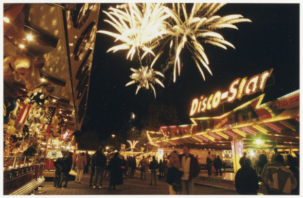 Eine Menschenmenge steht vor einer Karussellfahrt in der Nacht, beleuchtet von Lichtern und umgeben von Gebäuden mit Namensschildern, mit Bäumen und Feuerwerk im Hintergrund.