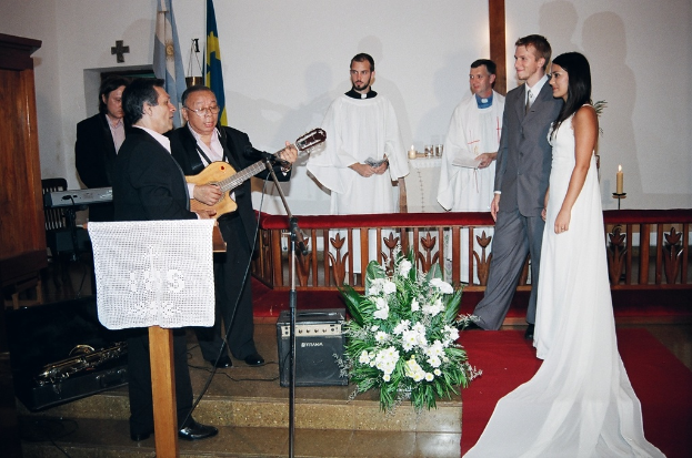 Eine HochzeitsSzene in einem Raum mit zwei Pastoren, einer Braut, einem Bräutigam und drei Musikern; ein Podium mit Stoff vor den Musikern, ein Tisch mit Kerzen hinter den Pastoren, eine Kerze in einem Halter hinter dem Paar, ein Bukett, Lautsprecher, Taschen und roter Teppich auf dem Boden sowie Flaggen, eine Wand, eine Vitrine, einen Stuhl und ein Kreuz an der Wand im Hintergrund.