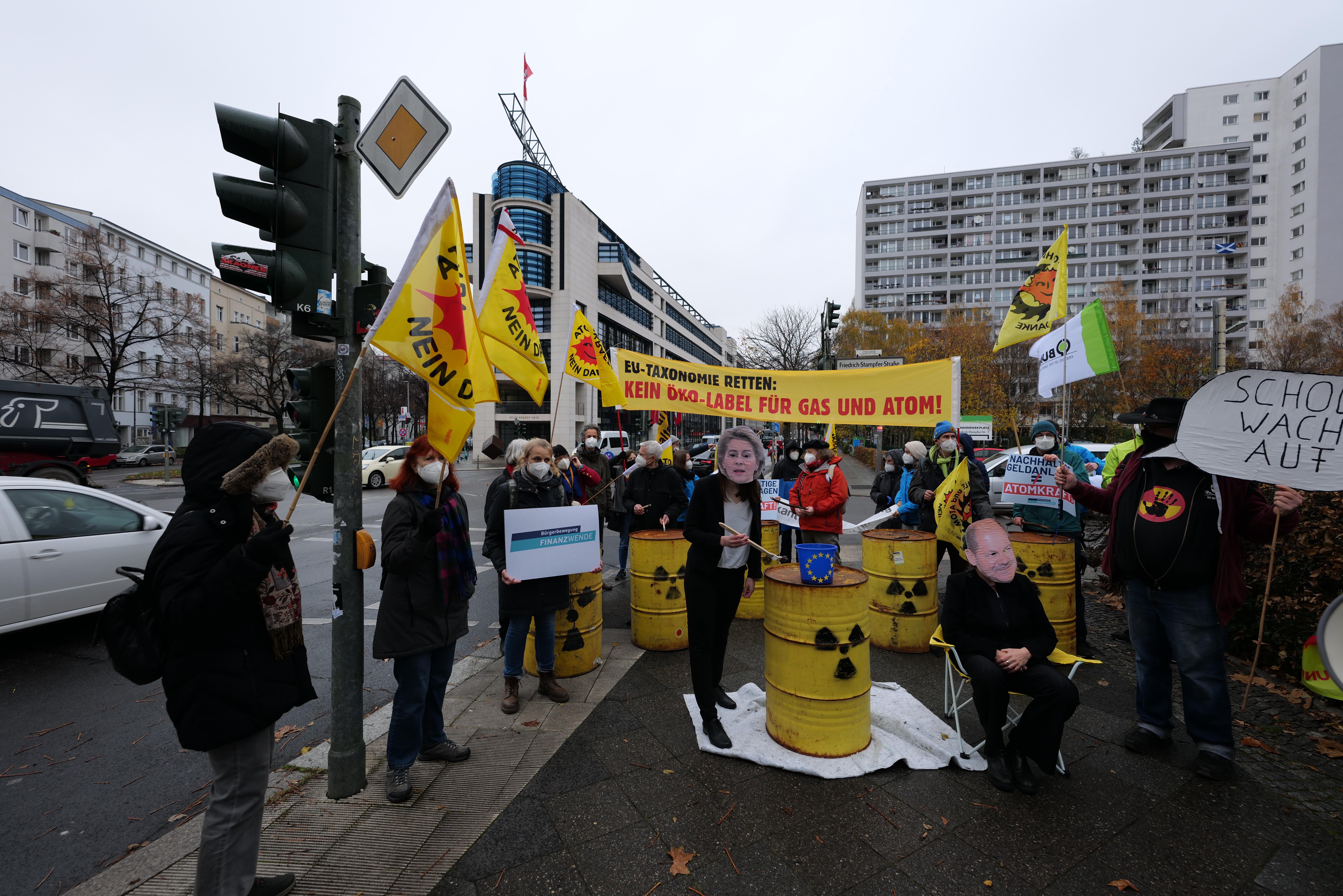 Eine Gruppe von Menschen steht auf einer Straße und hält gelbe Fässer und Schilder hoch, mit einer Person im Vordergrund, Fahrzeugen, Verkehrszeichen, Schautafeln, Bäumen, Gebäuden und einem klaren blauen Himmel im Hintergrund, die gegen Atommüll protestieren.