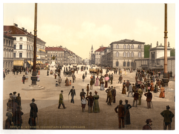 Ein altes Foto einer belebten Stadtstraße in München, Deutschland in den frühen 1900er Jahren, mit Menschen, die spazieren gehen, einige mit Schirmen, und einem Pferdewagen im Vordergrund, Gebäuden, Bäumen und Pfählen im Hintergrund und Text am unteren Rand des Bildes.