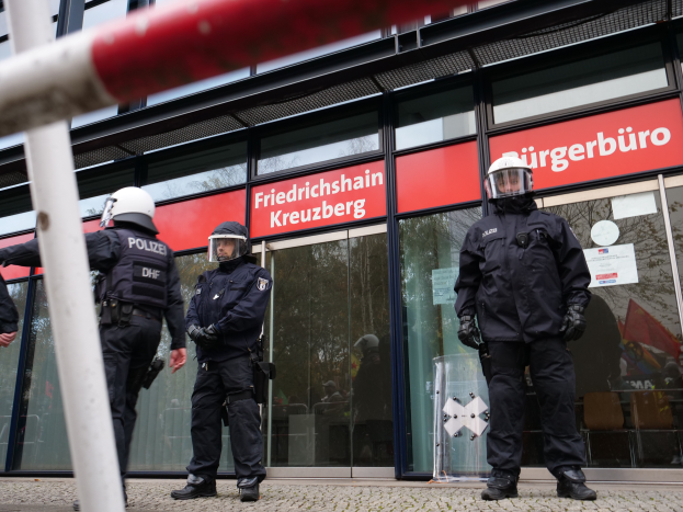 Eine Gruppe von Polizisten in Helmen und Handschuhen steht vor einem gläsernen Gebäude mit angeklebten Papieren, in der Nähe eines Pfahls, mit "Friedrichshain Kreuzberg"-Schildern im Hintergrund.