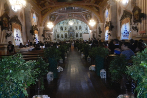 Eine Hochzeitszeremonie in einer Kirche mit Sitzplätzen und Stehplätzen, dekoriert mit Zimmerpflanzen entlang des Gangs, Skulpturen an den Wänden, Vorhängen an den Fenstern, hängenden Kronleuchtern und einem Bildschirm im Hintergrund.