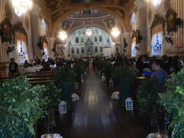 Eine Hochzeitszeremonie in einer Kirche mit Sitzplätzen und Stehplätzen, dekoriert mit Zimmerpflanzen entlang des Gangs, Skulpturen an den Wänden, Vorhängen an den Fenstern, hängenden Kronleuchtern und einem Bildschirm im Hintergrund.
