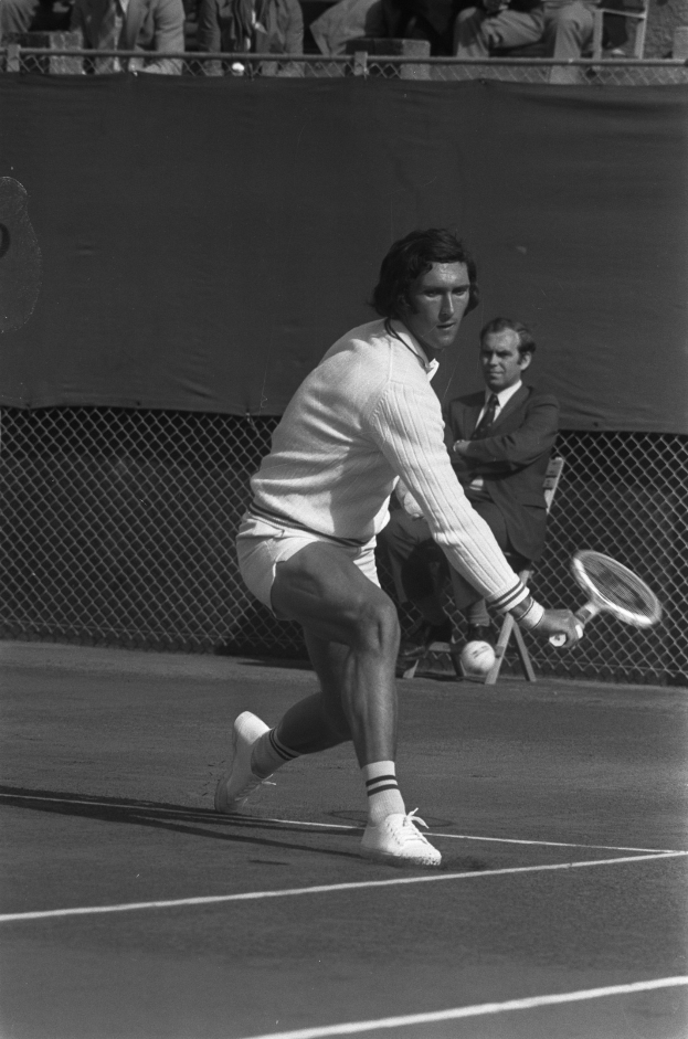 Ein professioneller Tennisspieler in Schuhen schwingt einen Schläger gegen einen Ball während eines Matches, mit Netz und Zuschauern im Hintergrund.