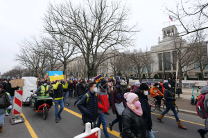 Eine große Gruppe von Menschen nimmt an einer Protestdemo auf einer Straße in Washington, D.C. teil, wobei einige Schilder und Banner halten und andere Fahrräder fahren, sowie Schilder, Bäume und ein klarer blauer Himmel im Hintergrund zu sehen sind.