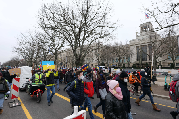 Eine große Gruppe von Menschen nimmt an einer Protestdemo auf einer Straße in Washington, D.C. teil, wobei einige Schilder und Banner halten und andere Fahrräder fahren, sowie Schilder, Bäume und ein klarer blauer Himmel im Hintergrund zu sehen sind.
