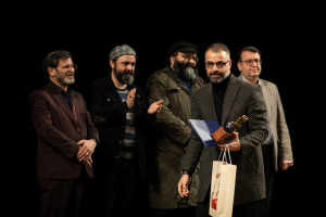 Eine Gruppe von Männern steht auf einer Bühne mit einem, der eine Tasche und eine Trophäe hält, feiernd die Eröffnung der Berliner Filmfestspiele gegen einen dunklen Hintergrund.