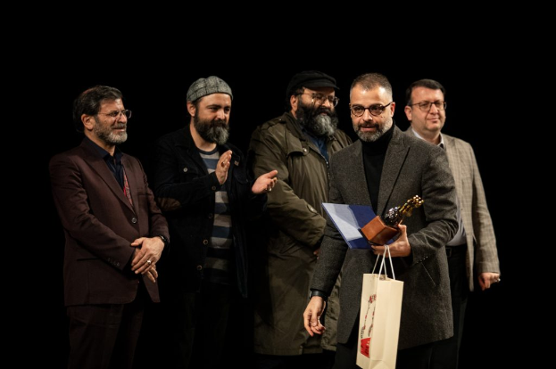 Eine Gruppe von Männern steht auf einer Bühne mit einem, der eine Tasche und eine Trophäe hält, feiernd die Eröffnung der Berliner Filmfestspiele gegen einen dunklen Hintergrund.