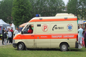 Ein Rettungswagen steht auf einem Rasenfeld neben einer Menschenmenge, einige tragen Mützen, mit Zelten, Bäumen, Gebäuden und einem klaren blauen Himmel im Hintergrund.