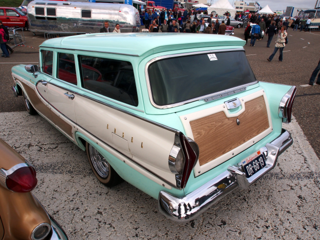 Ein gr├╝ner und wei├čer Kombiwagen auf einem vollen Parkplatz mit Zelten, Masten, B├Ąumen und einem klaren blauen Himmel im Hintergrund.