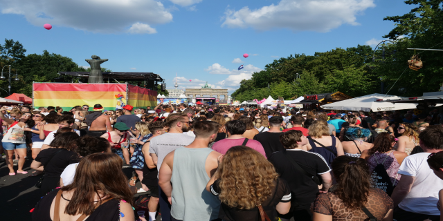Eine große Menschenmenge, die eine Straße mit Zelten, Bäumen, Pfählen, Lichtern und einer Statue gesäumt, mit Gebäuden im Hintergrund und einem bewölkten Himmel mit Ballons bei der Christopher Street Day Parade in Berlin.