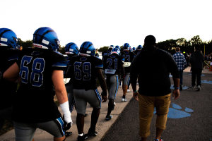 Gruppe von Füßballspielern in Helmen und Stiefeln, die eine Straße mit Bäumen und einem klaren blauen Himmel im Hintergrund entlanggehen.