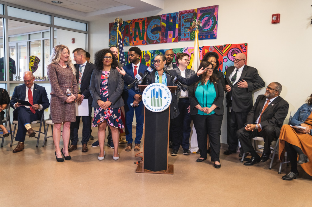 Eine Gruppe von Menschen steht vor einem Podium mit einem Mikrofon und einer Tafel mit Text, mit sitzenden und stehenden Teilnehmern in einem Raum mit Flaggen, einem Feuerlöscher und Deckenleuchten.