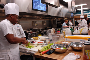 Eine Gruppe von Köchen in einer Küche, die Essen auf einem Schneidbrett zubereiten, mit einem Koch im Vordergrund in einer weißen Schürze und Mütze, der ein Messer hält und von verschiedenen Utensilien, Schüsseln, Gemüse, einem Handy, Flaschen und anderen Gegenständen umgeben ist, mit einem Fernseher und Deckenleuchten im Hintergrund.