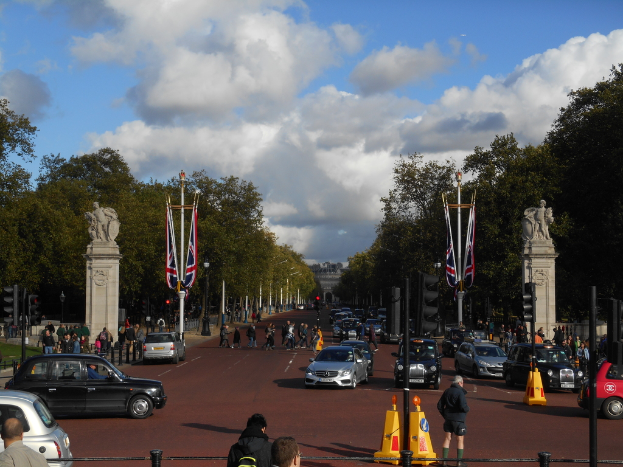 Ein Denkmal steht auf einer Straße mit wenigen fahrenden Autos und Fußgängern und ist von Verkehrsampeln, Pfählen und Fahnen umgeben. Im Hintergrund sind Bäume und Gebäude zu sehen, der Himmel ist bewölkt.