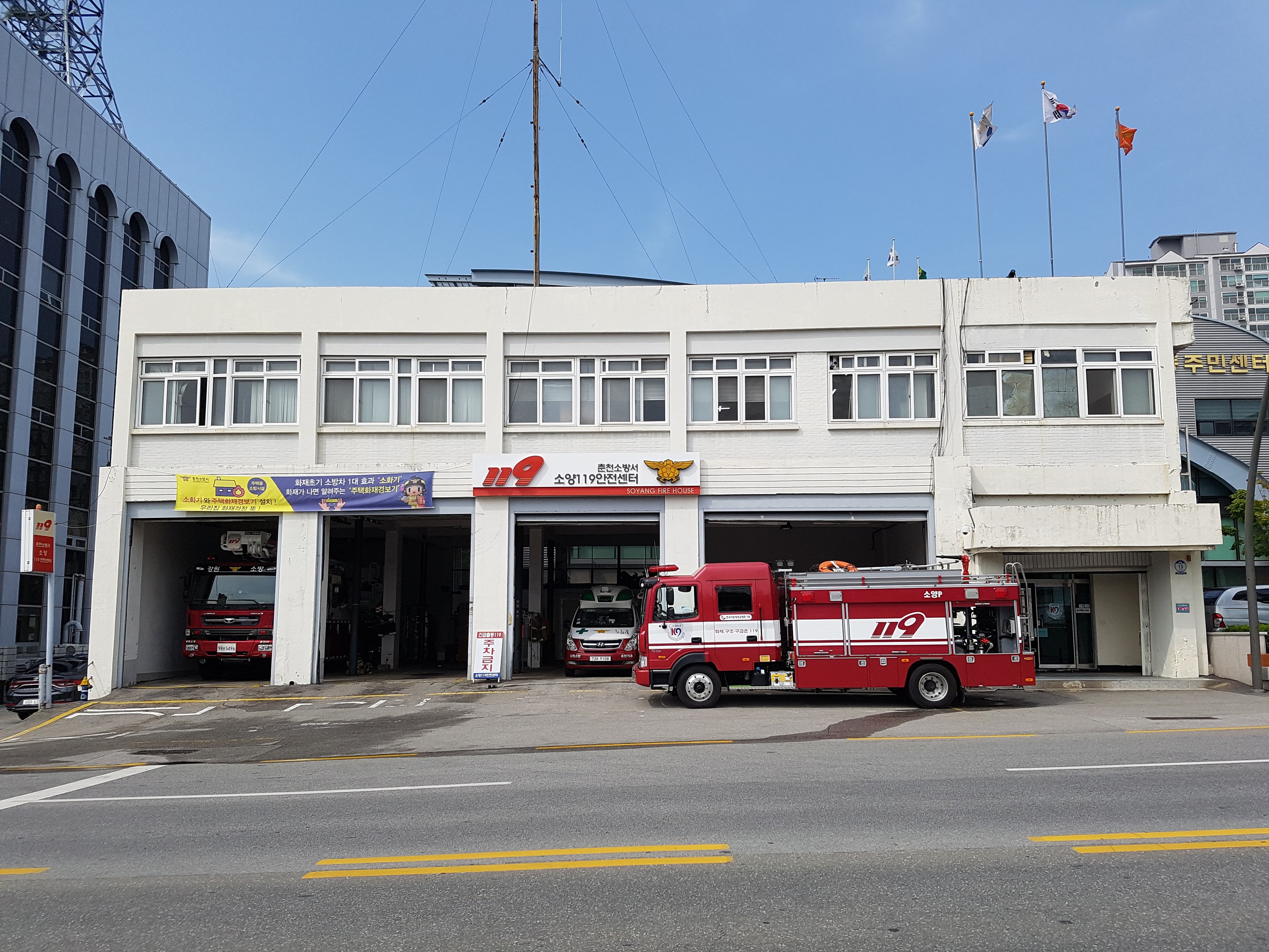 Feuerwache mit mehreren Feuerwehrautos vor dem Gebäude, das Fenster, Banner und Fahnen aufweist, unter einem bewölkten Himmel mit einem Turm links.