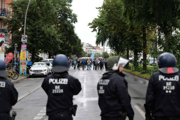 Eine Gruppe von Polizisten in schwarzen Uniformen und Helmen steht an der Straße, mit Menschen, Fahrzeugen, Fahrrädern, Pfosten, Plakaten, Bäumen, Gebäuden und einer Banner mit Text im Hintergrund.
