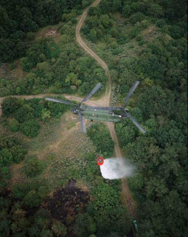 Hubschrauber fliegt über einen dichten grünen Wald mit einer Straße im Hintergrund, Text am unteren Bildrand.
