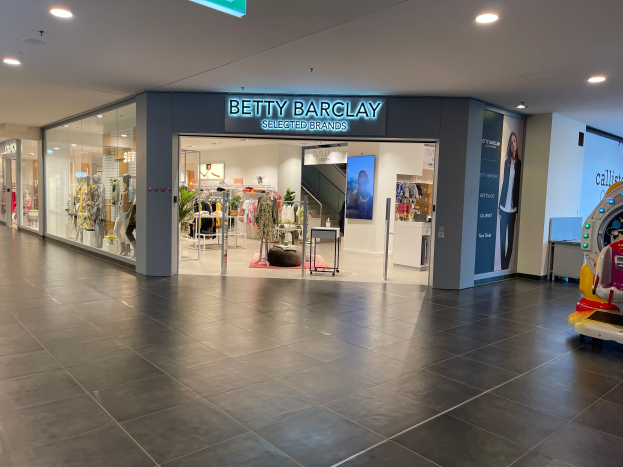 Das Innere eines Betty Barclay-Ladens in einem Einkaufszentrum, mit Schaufensterpuppen in Kleidern, hängenden Kleidern, Tischen, Pflanzen und anderen Gegenständen, sowie einem Spielzeugfahrzeug auf der rechten Seite und einer Tafel mit der Aufschrift "Betty Barclay Selected Brands" oben.