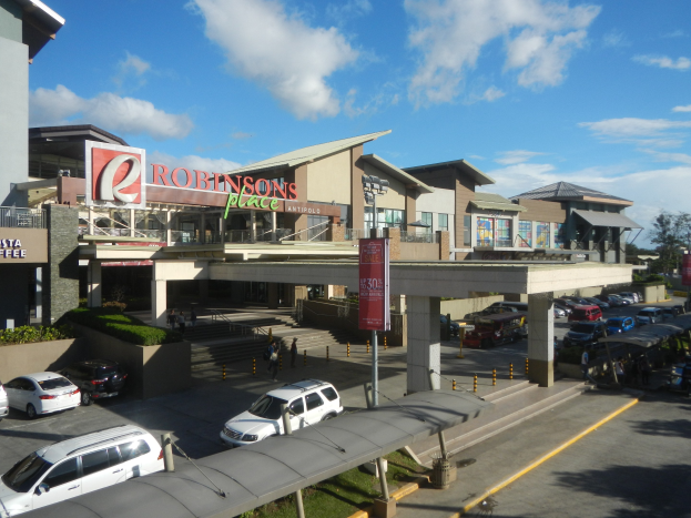 Ein belebter Einkaufszentrum mit Gebäuden, Geschäften, Kraftfahrzeugen, Fußgängern, Straßenmöblierung, Grünflächen und einem bewölkten Himmel.