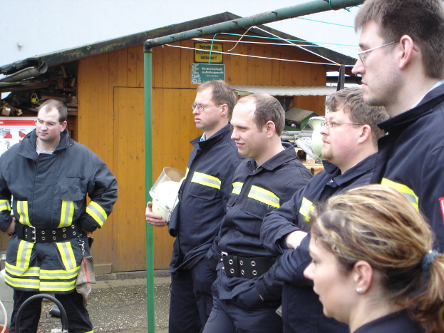 Eine Gruppe von Männern in blauen Feuerrettungsuniformen steht links und schaut auf ein kleines hölzernes Schuppenhaus und Rohrrahmen.