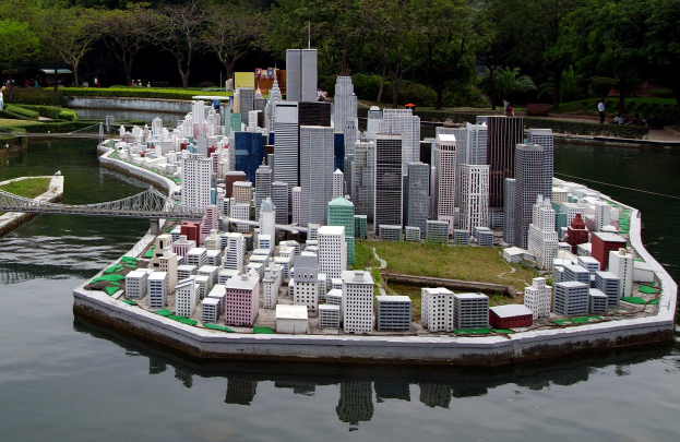 Ein detailliertes Miniaturmodell einer Stadtlandschaft auf Wasser, mit Gebäuden, Grünflächen, einer Brücke, Menschen, Pflanzen, Bäumen und einem Himmelhintergrund.