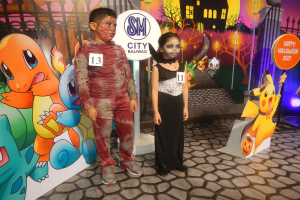 Zwei Kinder in Halloween-Kostümen stehen nebeneinander vor einer Wand mit einer Painting von einem Gebäude, Bäumen, Laternenmasten und einem Zaun sowie Schildern mit Text und Tierbildern und festlicher Beleuchtung.