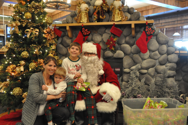 Eine Frau, Kinder und ein als Santa Claus verkleideter Mann mit Brille und Handschuhen stehen in der Nähe eines geschmückten Weihnachtsbaums, einer Wand mit Felsen, Spielzeugen, Dekorationen und einem Korb mit Gegenständen, mit Lichtern im Hintergrund.
