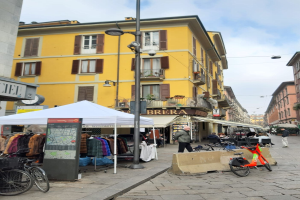 Eine belebte Stadtstraße mit Fußgängern, parkenden Fahrrädern, aufgehängter Wäsche, einem Zelt, Straßenmöblierung, Topfpflanzen und einem bewölkten Himmel.